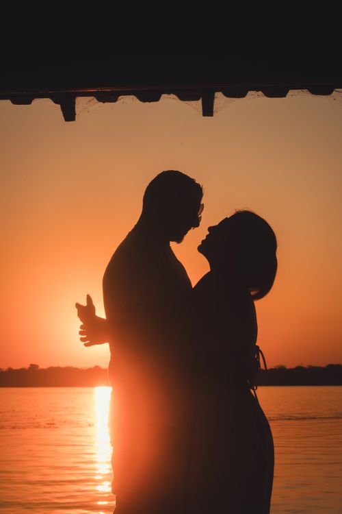 Free Silhouetted couple embraces at sunset, creating a romantic scene by the lake under an orange sky. Stock Photo