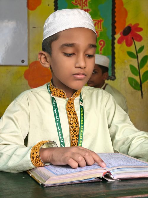 Free Young boy in traditional clothing reading a book in a classroom setting. Stock Photo