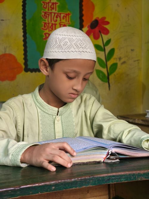 Free A young boy in traditional attire reads a book at a desk indoors, focusing intently. Stock Photo