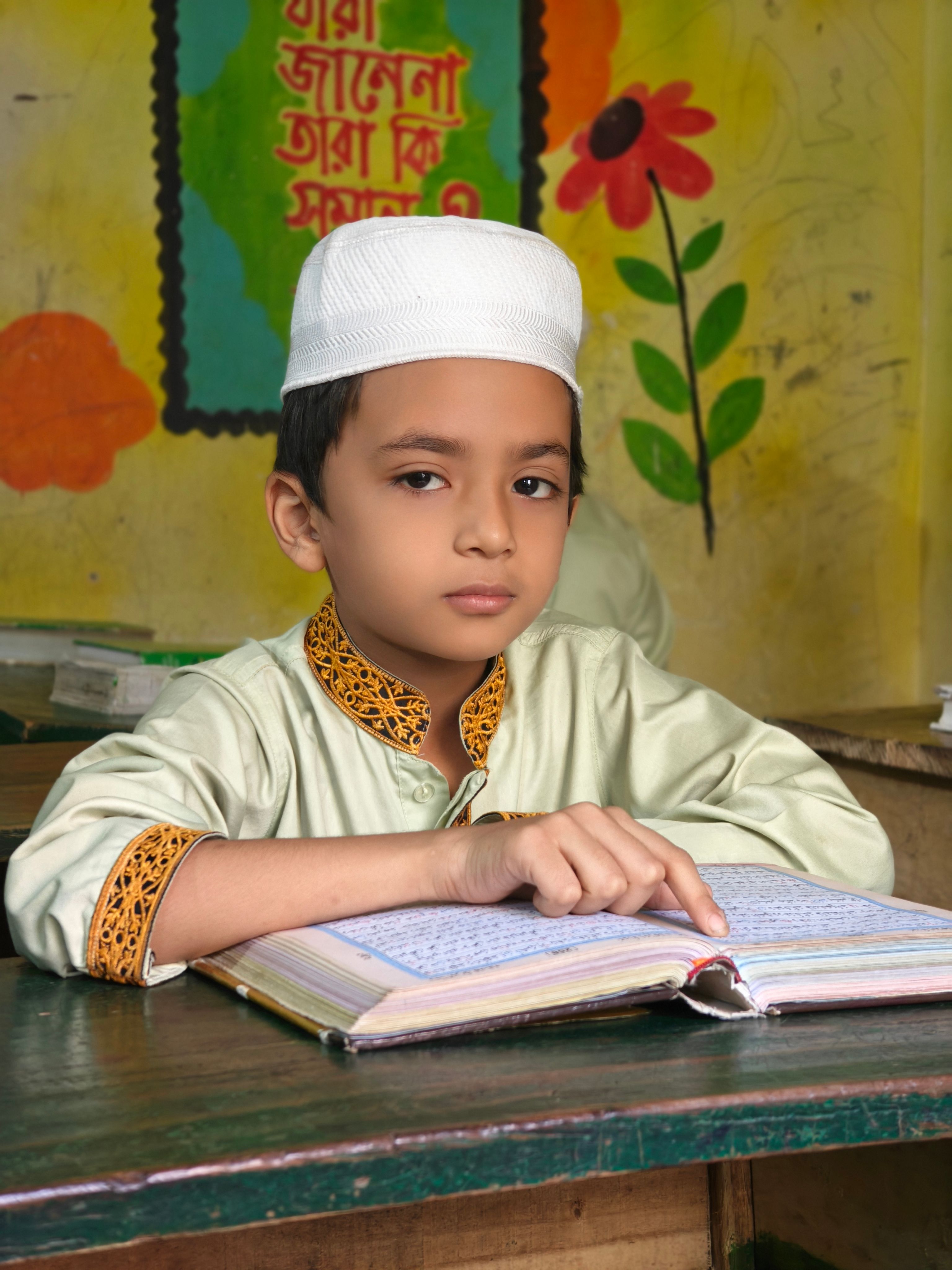 A young boy in traditional attire reading the Quran inside a madrasa classroom.