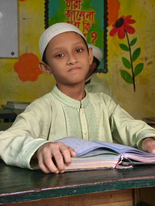 Free Young boy in traditional dress reading a textbook in a colorful classroom setting. Stock Photo