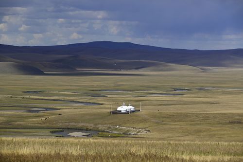 Free A Mongolian ger sits alone on expansive grasslands under a dramatic sky, surrounded by hills. Stock Photo