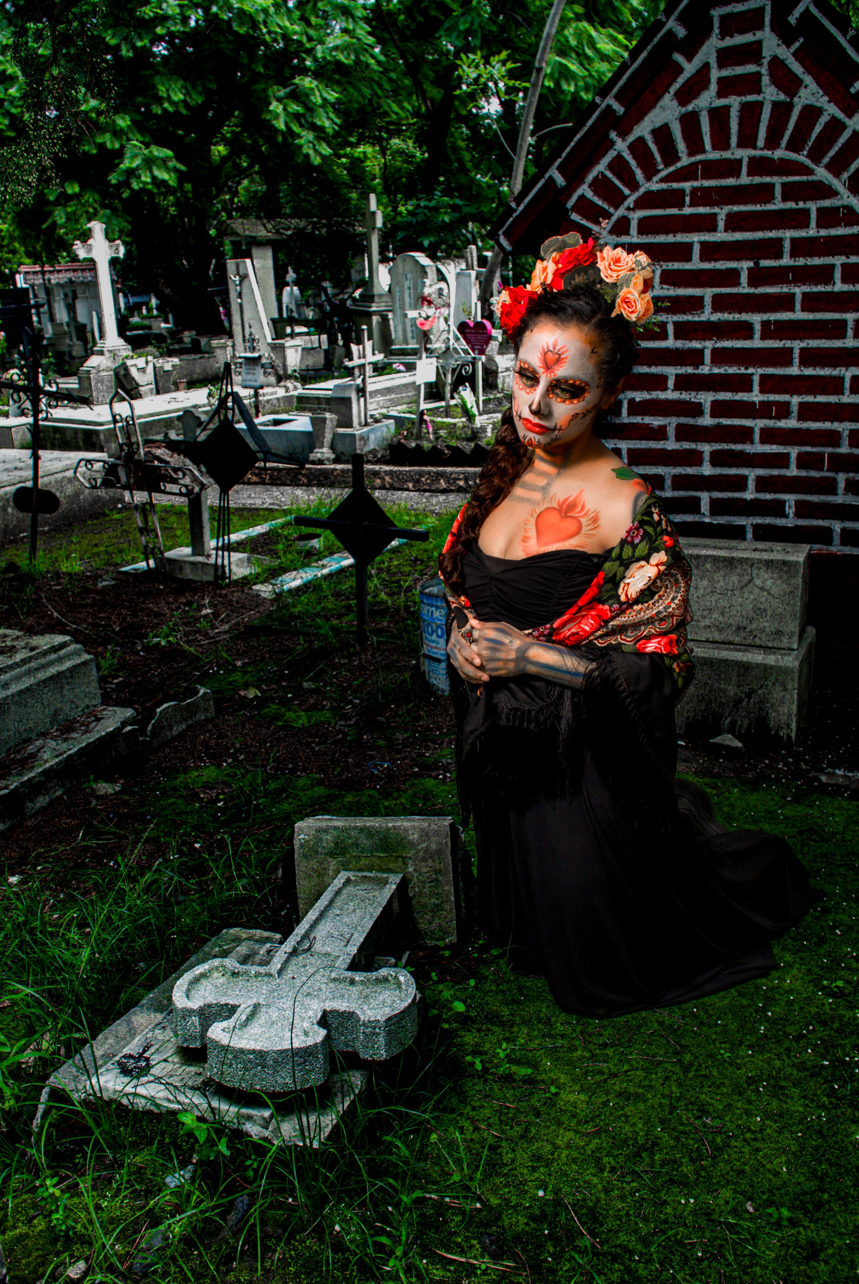 Woman in Day of the Dead makeup and attire kneels in a cemetery, evoking cultural tradition.