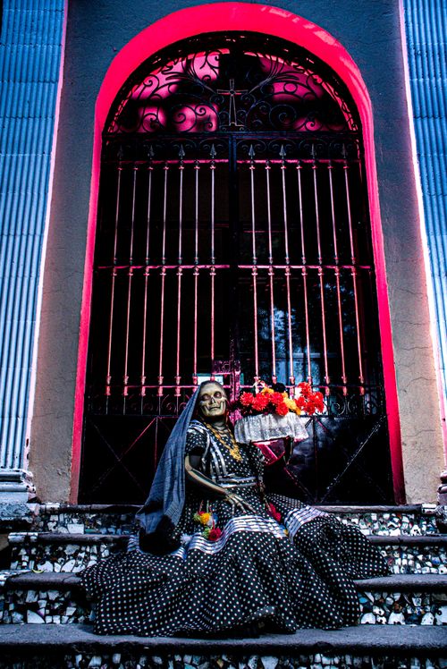 Free Catrina figurine dressed in traditional attire with flowers, set against an ornate gate in vibrant lighting. Stock Photo