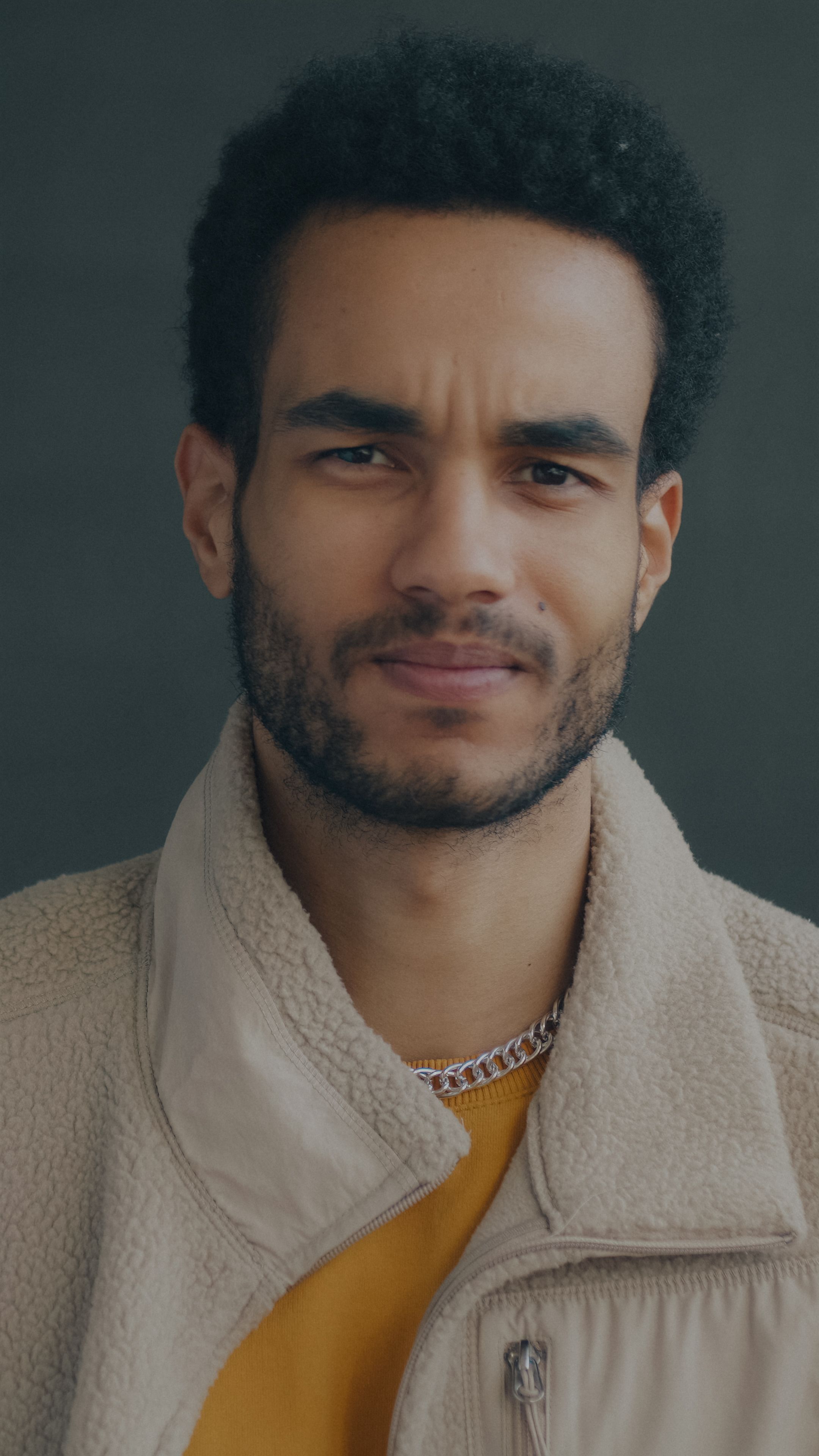 Close-up portrait of a confident young man wearing a beige fleece and yellow top against a dark background.