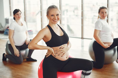 Adult pregnant females in activewear sitting on fit balls while doing exercises with instructor in modern studio of fitness center