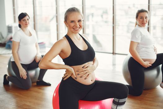 Adult pregnant females in activewear sitting on fit balls while doing exercises with instructor in modern studio of fitness center