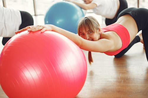 Slim young sportswoman doing stretching exercise with fitness ball together with crop pregnant women during workout in modern sport studio and looking at camera with smile