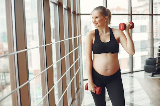 Young pregnant female in sportswear smiling and looking away while standing near glass wall in modern gym and training with red dumbbells