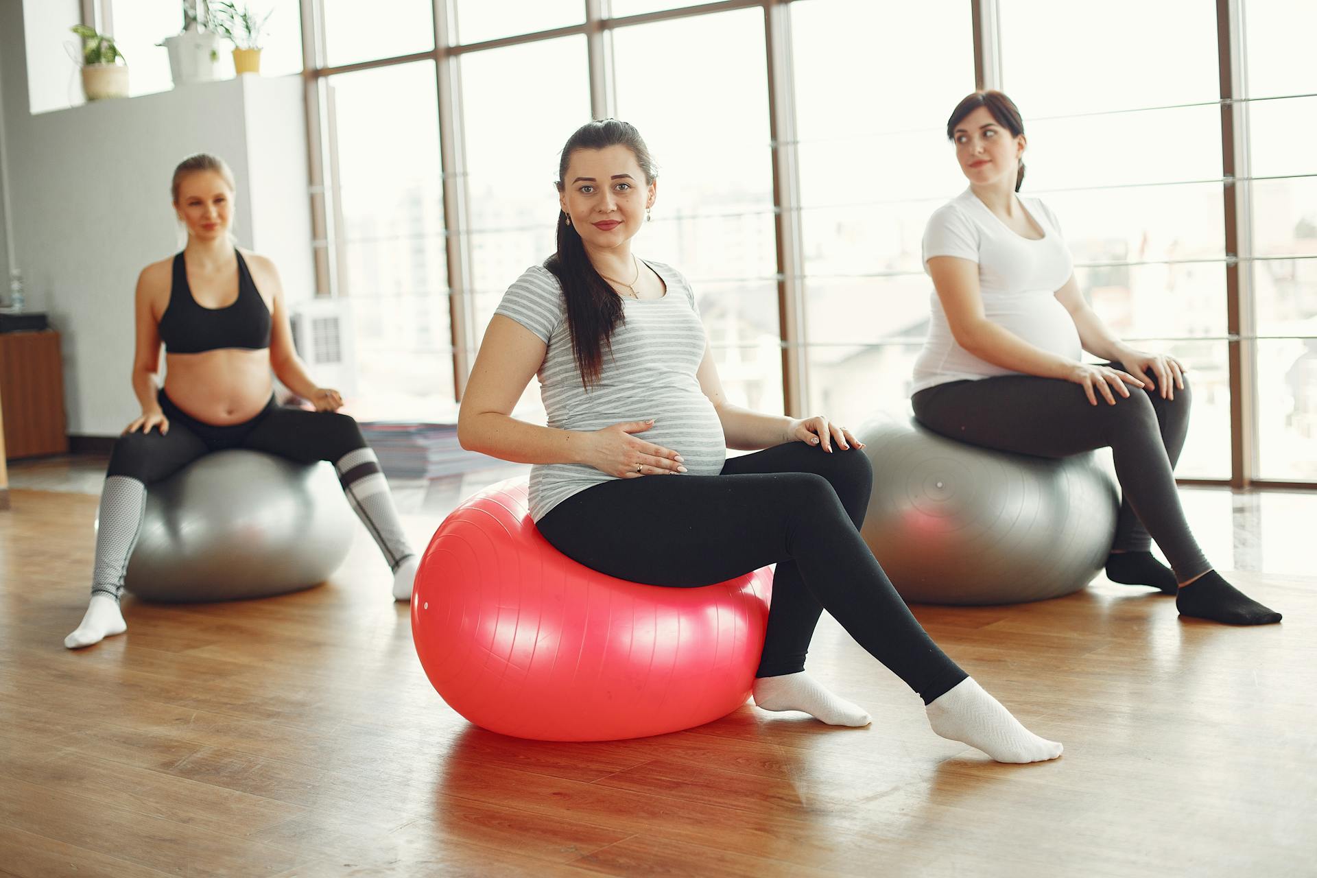 Calm pregnant ladies sitting on fit balls during prenatal training in spacious light gym class at daytime while caring about health