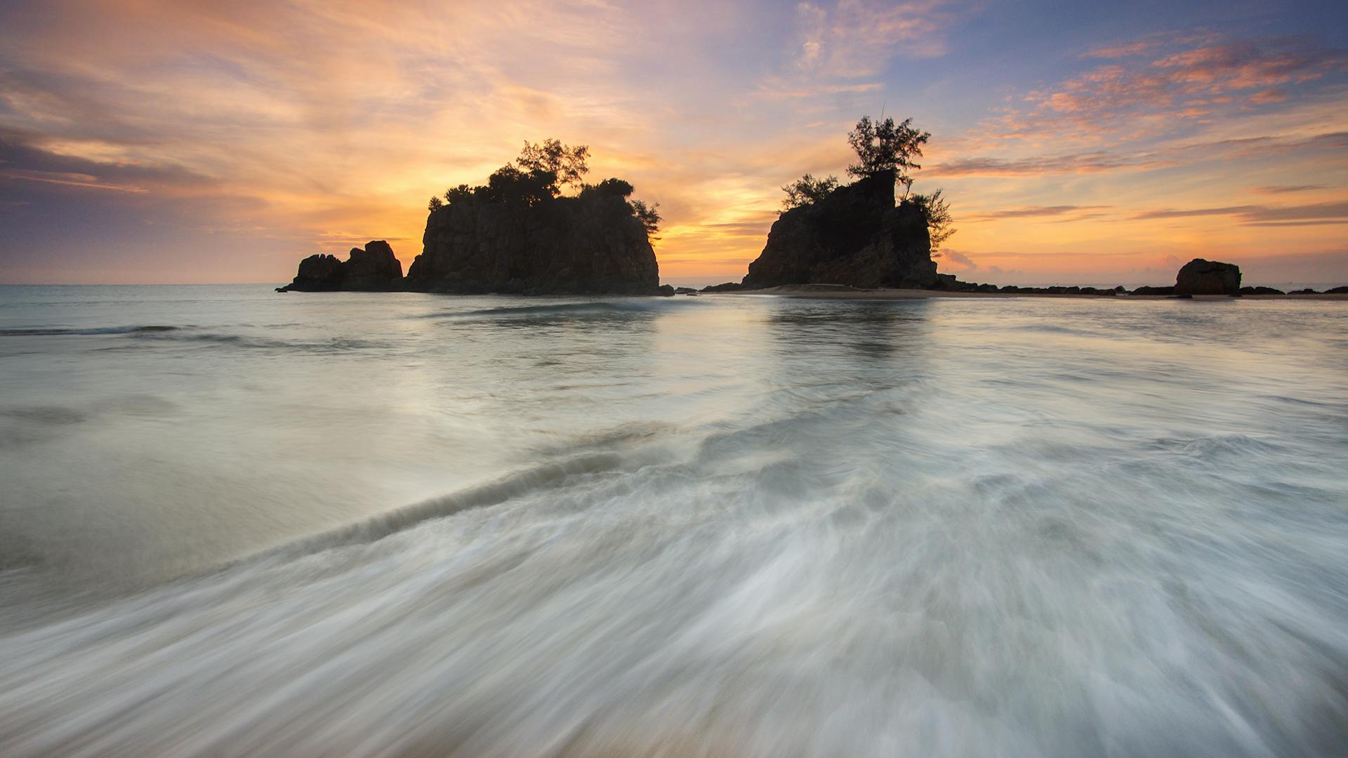 Tranquil ocean waves flow by rocky islands during a vibrant and colorful sunset.