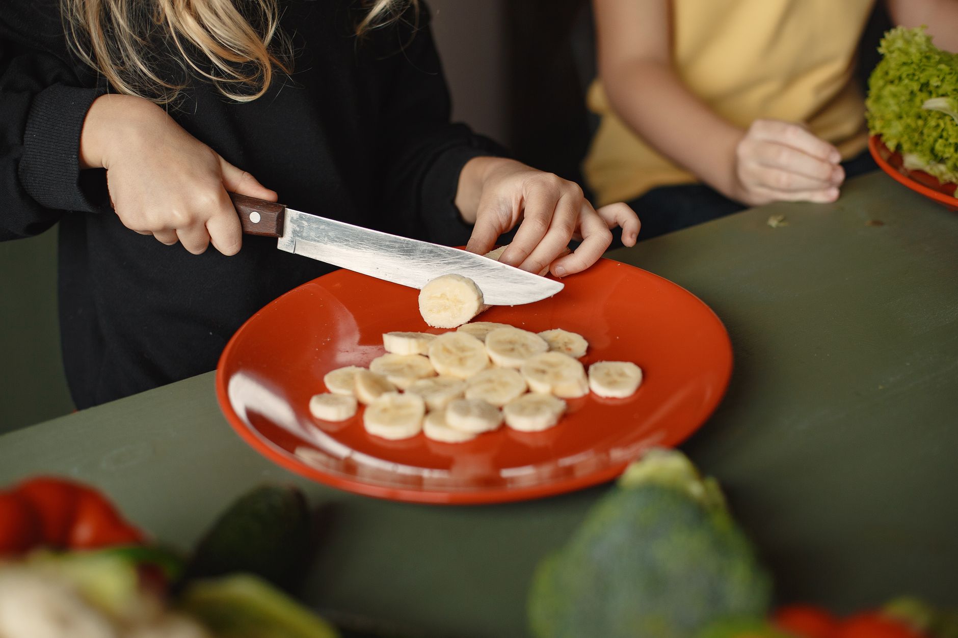 Two children preparing sliced bananas on a red plate in a kitchen setting.