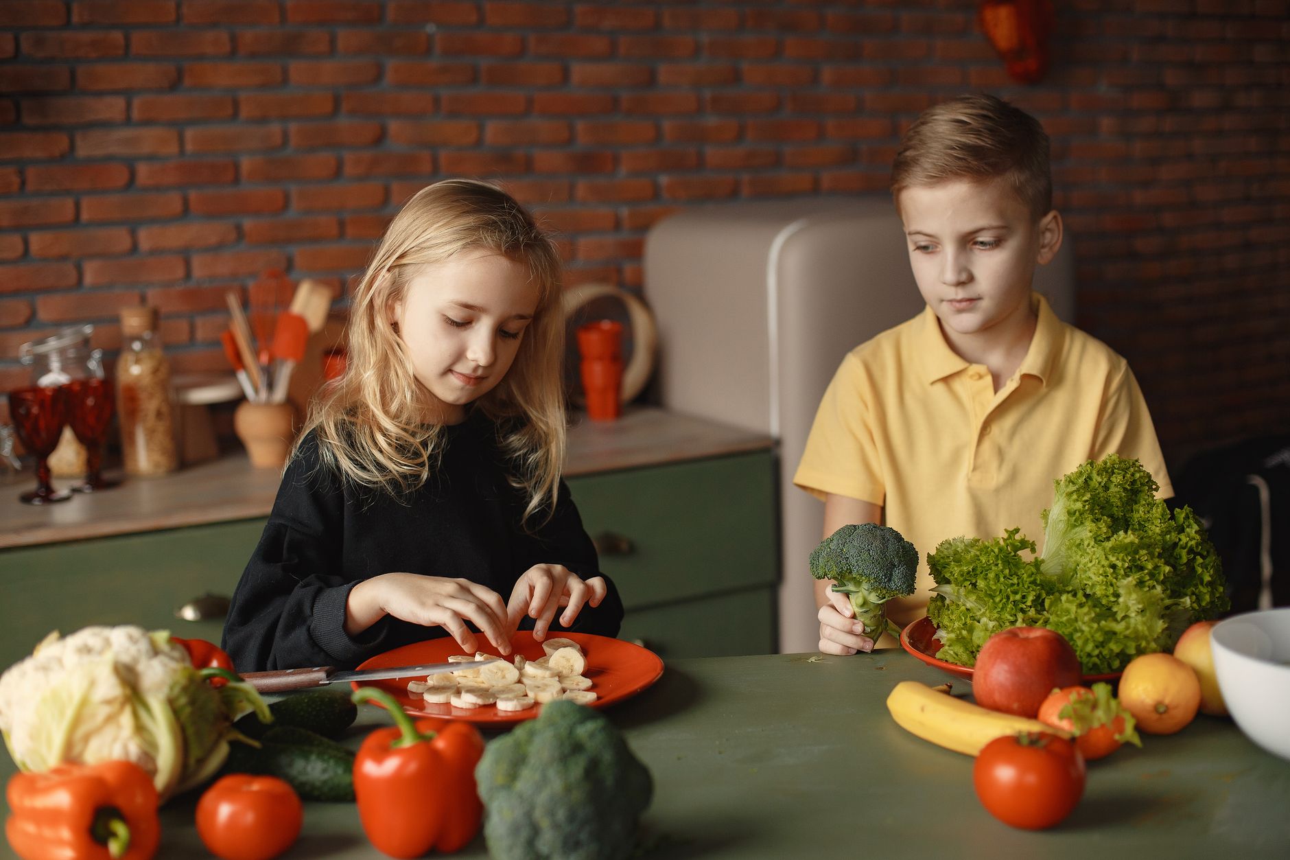 Positive little girl and boy sitting at desk with various fresh vegetables and fruits while preparing meal in loft kitchen with brick wall