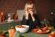 Funny cheerful girl looking through slice of pepper while preparing healthy vegetarian salad at table with plate with knife and salad bowl