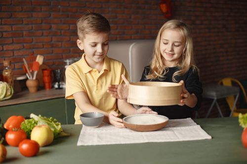 Free Content little friends in casual wear sifting flour through wooden sieve into round plate put on towel on table with fresh vegetables in loft style kitchen at home Stock Photo