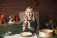 Happy little blond kid in casual wear playing with flour put into round wooden plate while standing near big sieve in kitchen behind brick wall