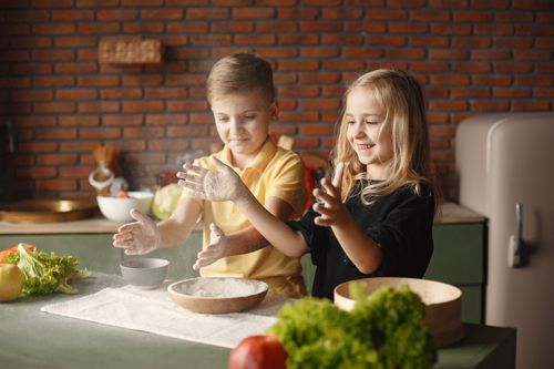Free Cheerful siblings in casual clothes playing with flour put into wooden plate on towel while standing near table with fresh vegetables in loft style kitchen in daylight Stock Photo