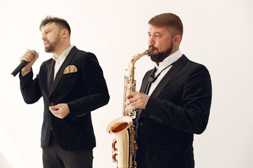 Free Talented male musician playing saxophone near inspired male vocalist singing beautiful song while standing together against white wall during celebration party or live music show Stock Photo