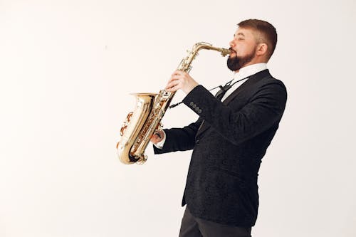 Side view of professional male musician wearing concert costume playing music on saxophone standing against white background in studio with passion and inspiration