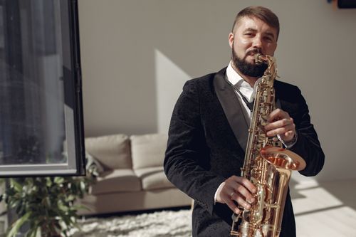 Free A man in formal attire plays saxophone in a well-lit room, creating a relaxed jazz ambiance. Stock Photo