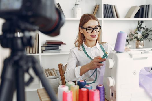 A fashion designer focuses on sewing with colorful threads in a modern studio with shelves and camera.