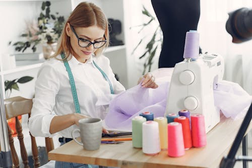 Woman in White Long Sleeve Shirt Wearing Black Framed Eyeglasses in Front of Her Sewing Machine