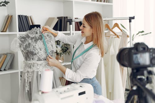 Talented young female tailor measuring wedding dress on mannequin while taking photos of designing process working in modern dressmaking atelier