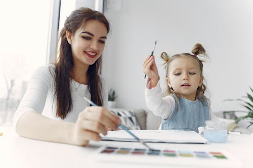 Woman in White Long Sleeve Shirt Holding Paint Brush Beside a Child in Blue Denim Dress Jacket