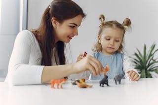 Adorable little girl sitting at white table with mother while playing with small toy wild animals for early education and development during spending time together at home