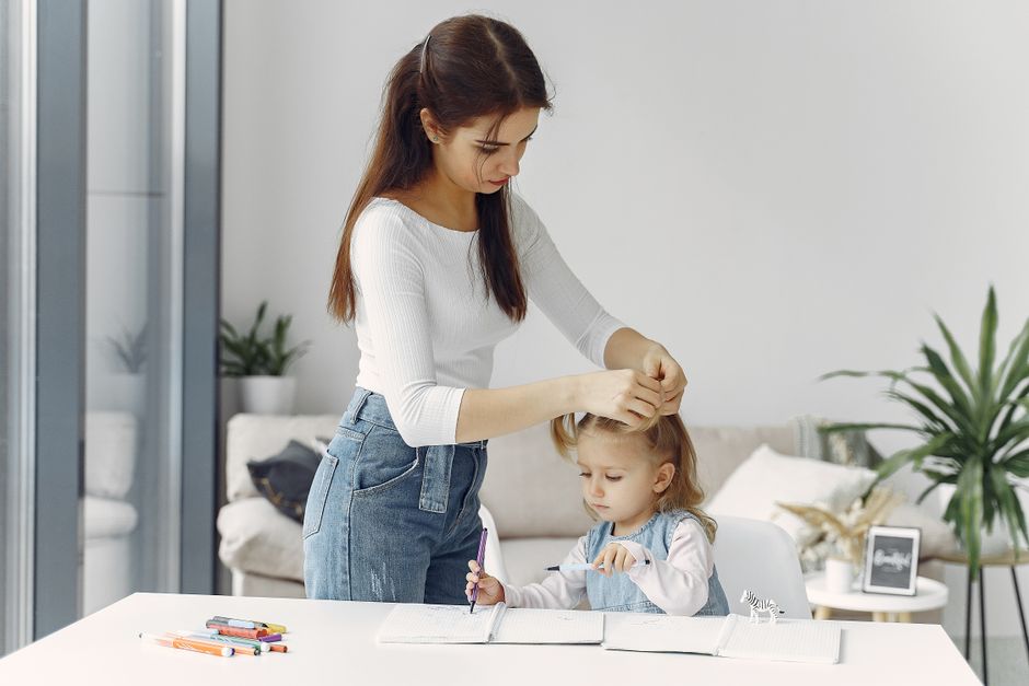 Una madre trenzando el cabello de su hija mientras la niña dibuja con lápices de colores en interiores.