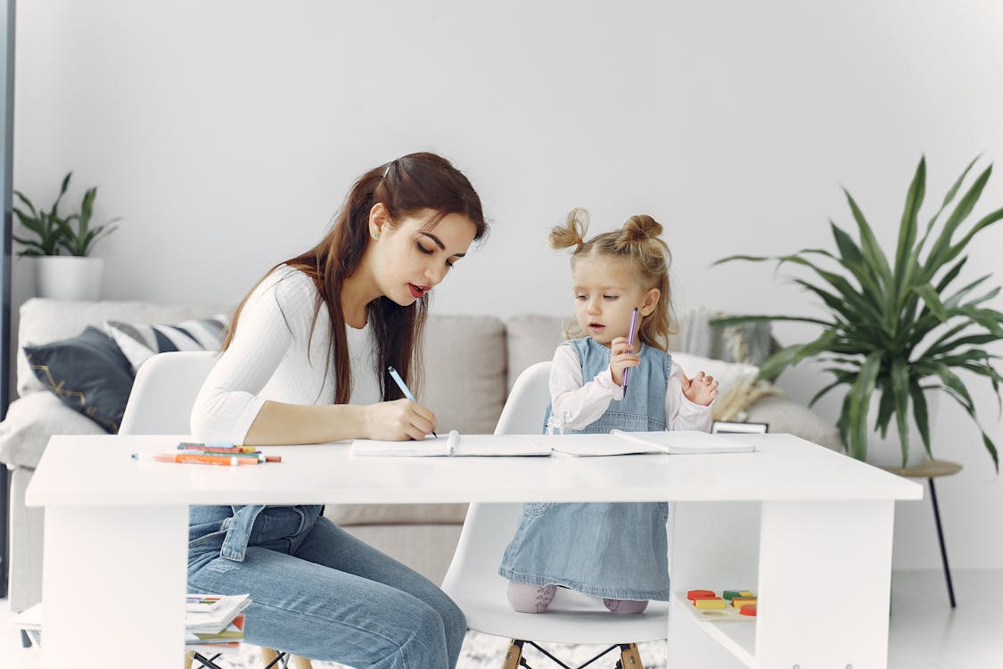 Free Woman in White Long Sleeve Shirt Sitting Beside Girl Writing on the Notebook Stock Photo
