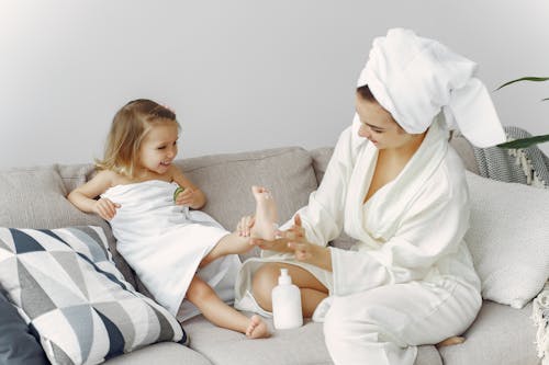 Woman in White Robe Sitting on Gray Couch Putting Lotion on Little Girls Foot