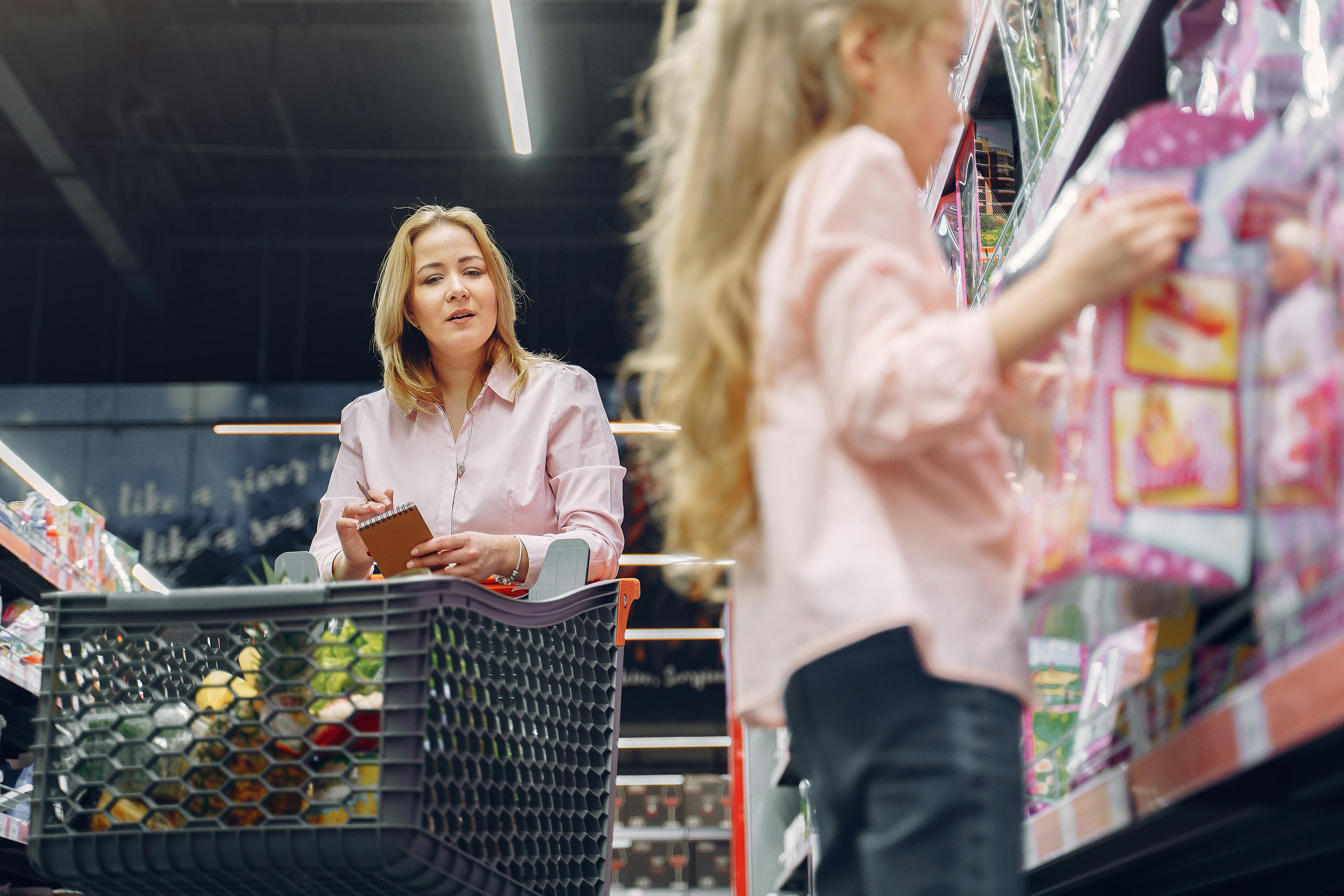 Woman pushing shopping cart through grocery store aisle with blurred child visible to the right, moving purposefully past product displays.