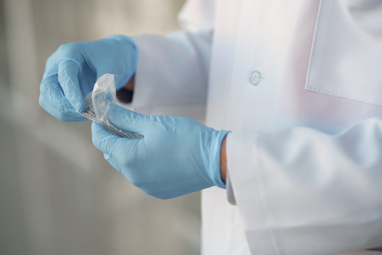 Person In Lab Gown Wearing Blue Gloves Opening Sterile Syringe