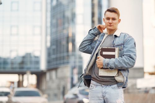 Free Young adult male student wearing denim jacket, standing confidently outdoors with notebooks. Stock Photo