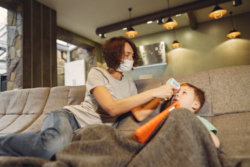 Low angle of slim mother in casual wear and face mask measuring boys body temperature using infrared thermometer while sitting on cozy sofa at home in afternoon during COVID 19