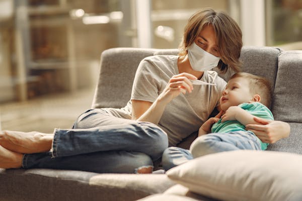 Mother in a mask checking her child’s temperature on the couch