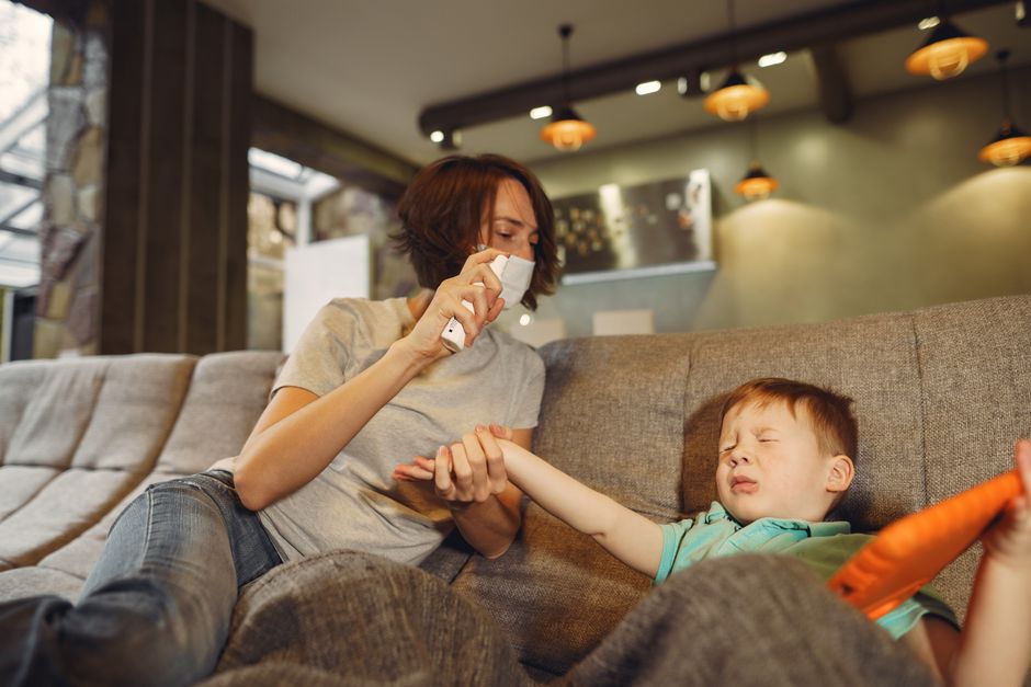 Photo by Gustavo Fring on Pexels Mother in face mask and casual wear taking medical spray and taking scared little boy hand with closed eyes and tablet while sitting on comfortable sofa at home during COVID 19 - Photo by Gustavo Fring on Pexels