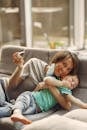 Happy mother in casual wear and downcast mask with medical spray embracing little boy while lying on sofa at home in afternoon during COVID 19 and looking at camera