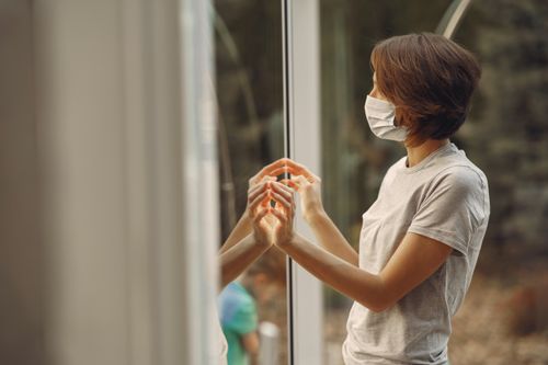 Free A woman in a face mask looks outside, her hands touching a glass window, reflecting in it. Stock Photo