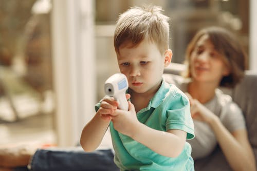 Adorable blond little child in casual clothes with infrared thermometer standing behind cheerful mom lying on sofa at home in daylight