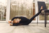 Positive female gymnast in sportswear stretching legs while sitting on transverse twine and putting one leg on chair near transparent door reflecting trees and looking at camera