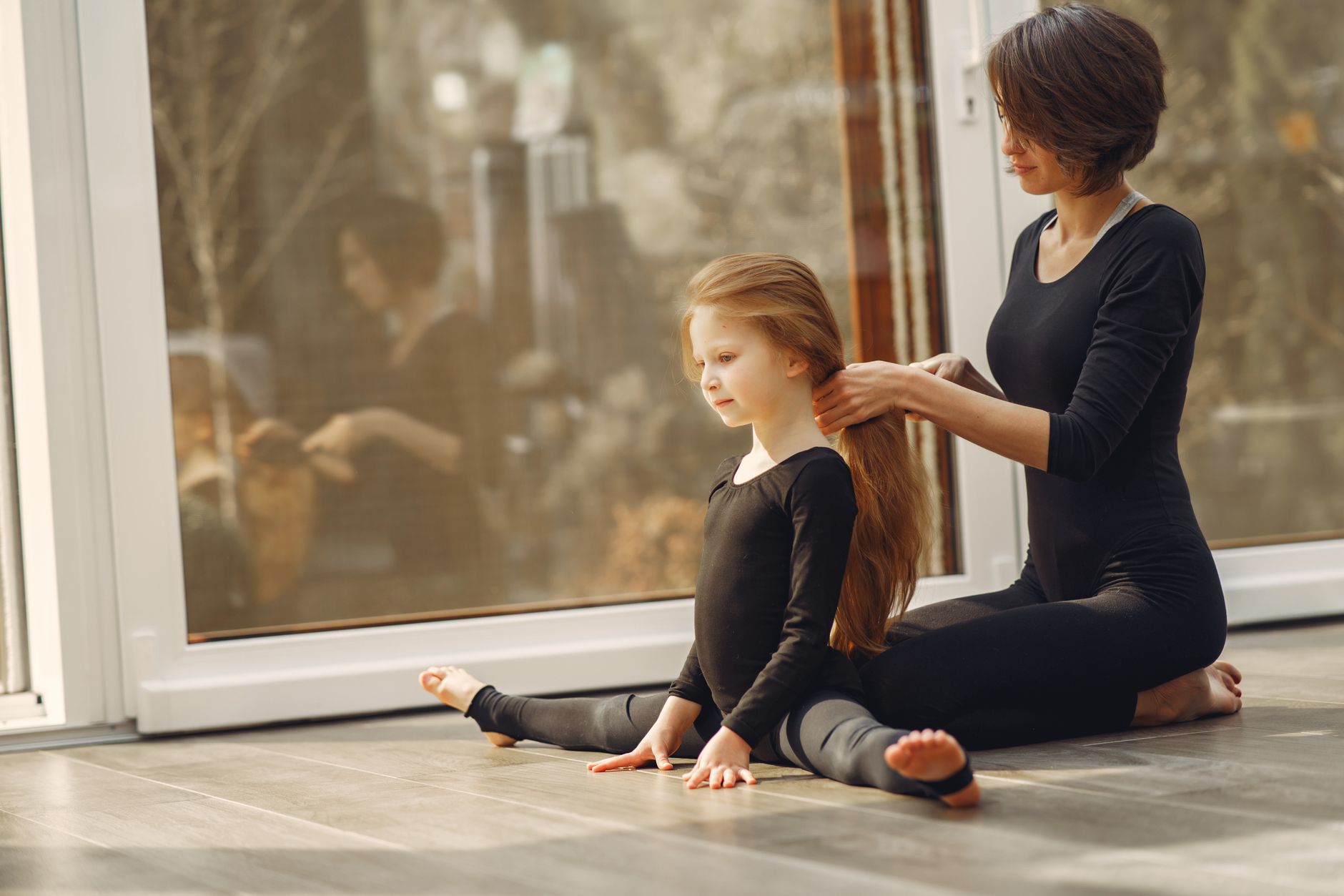 A mother and daughter in black outfits stretching at home, reflecting family bonding.