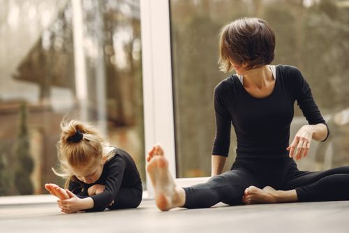 Free Anonymous female instructor in sportswear sitting near little blond gymnast doing tilt forward on floor near transparent door while stretching legs Stock Photo