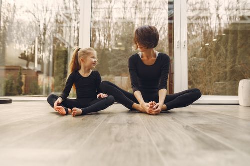 Free Anonymous mother in sportswear sitting near smiling little blond daughter with crossed legs on parquet while looking at each other in daylight Stock Photo