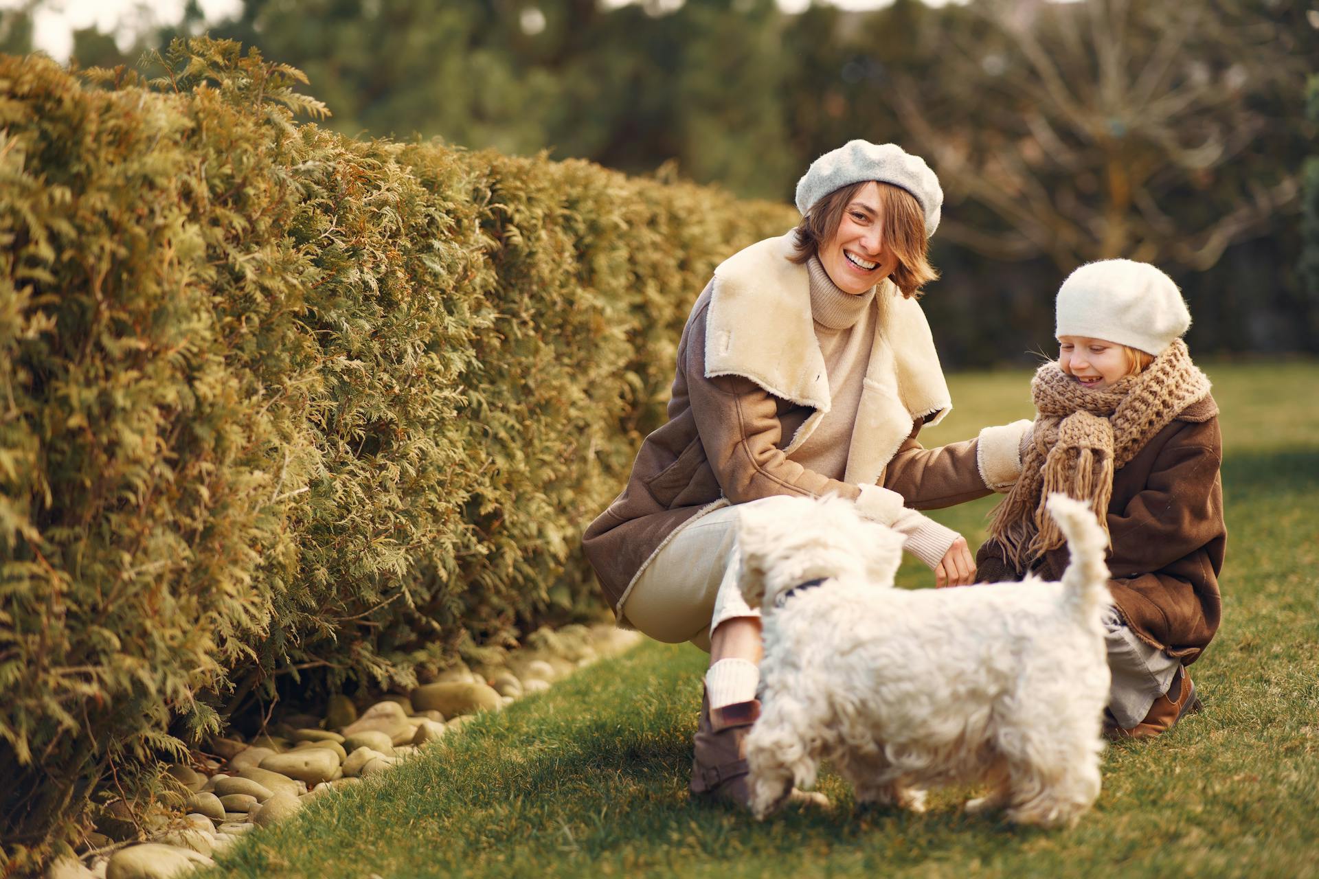 Une mère joyeuse en barette regarde la caméra et une petite fille souriante en vêtements chauds accroupie près d'un bichon frize chien sur l'herbe au soleil