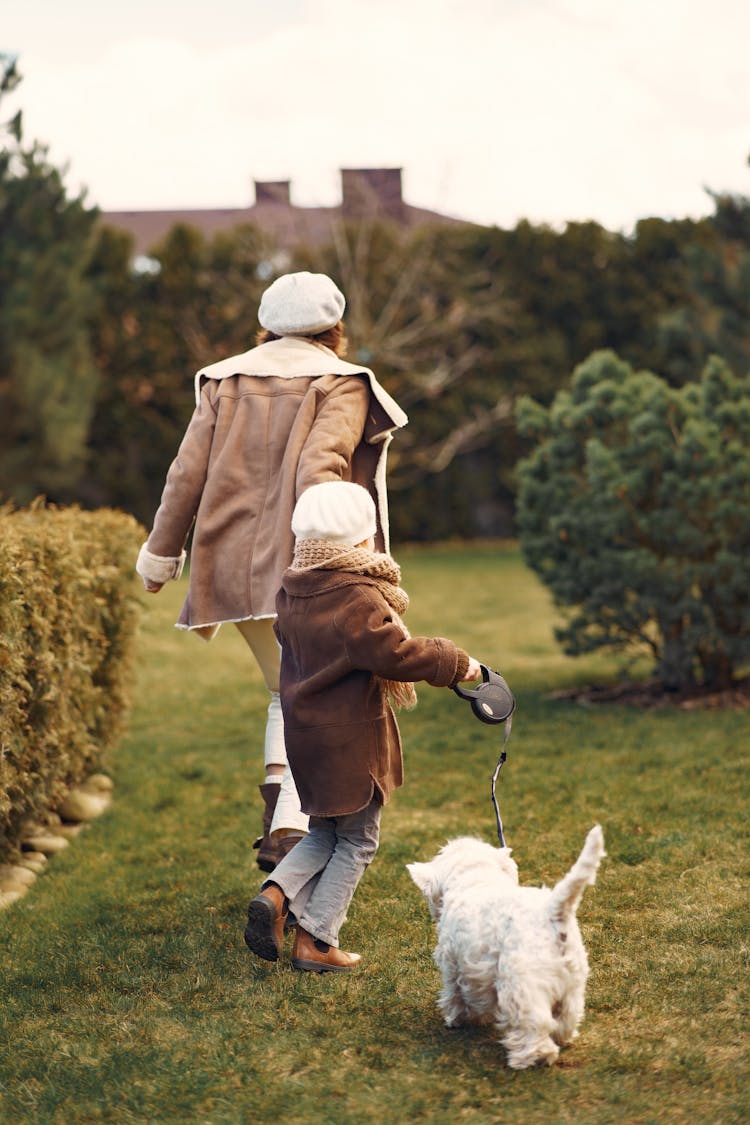 Unrecognizable Mother And Daughter Walking Bichon Frize Dog In Park