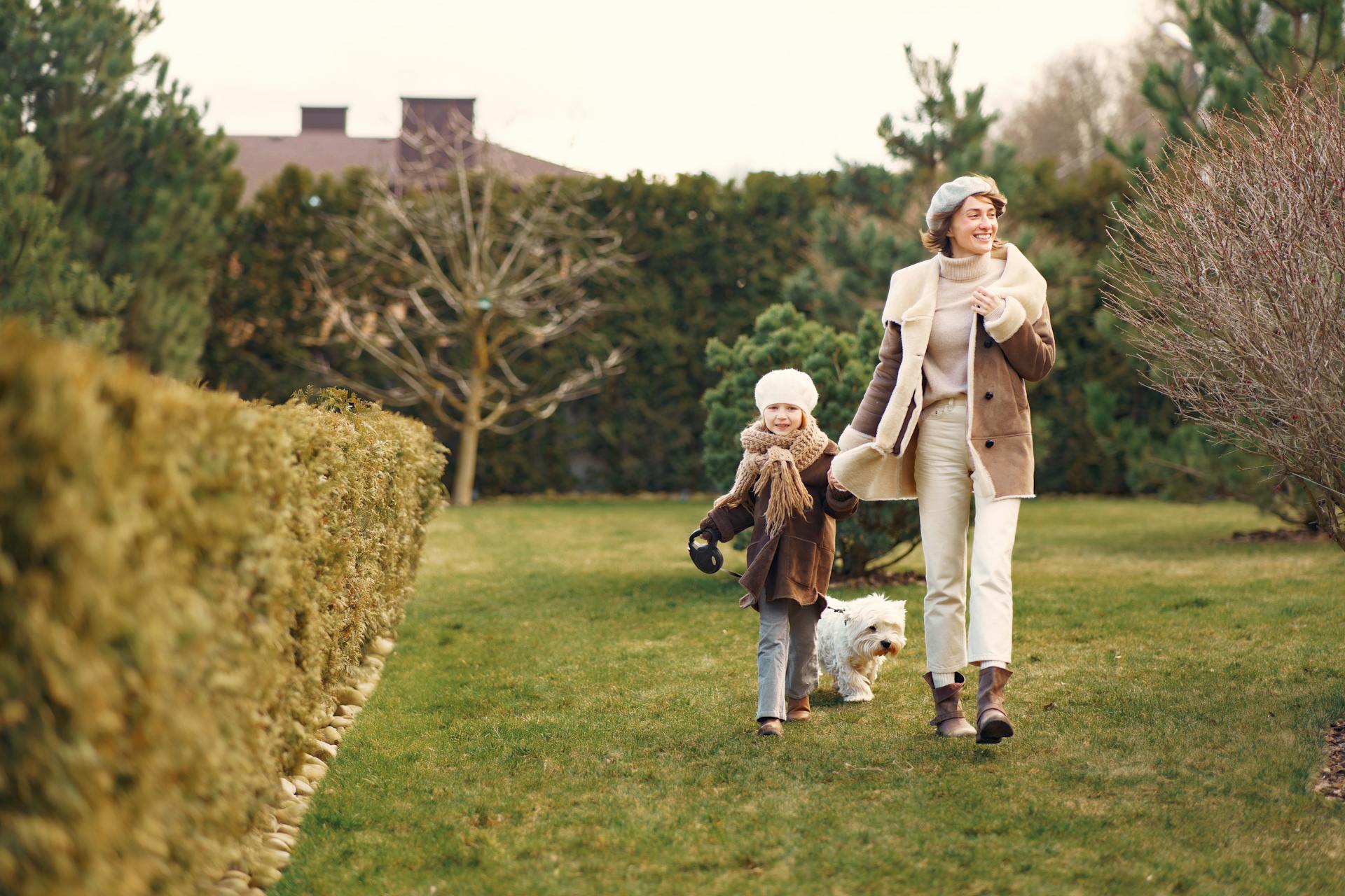 Cheerful mother and daughter in warm clothes walking little Bichon Frize dog in park near hedge and trees in daylight