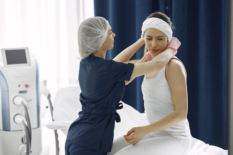 Woman In Blue Scrub Suit Helping Woman Sitting On Bed