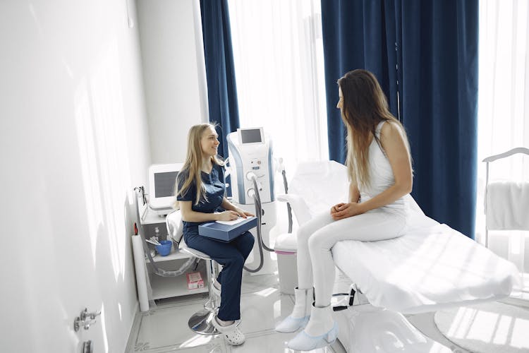 Woman In Blue Scrub Suit Helping Woman Sitting On Bed